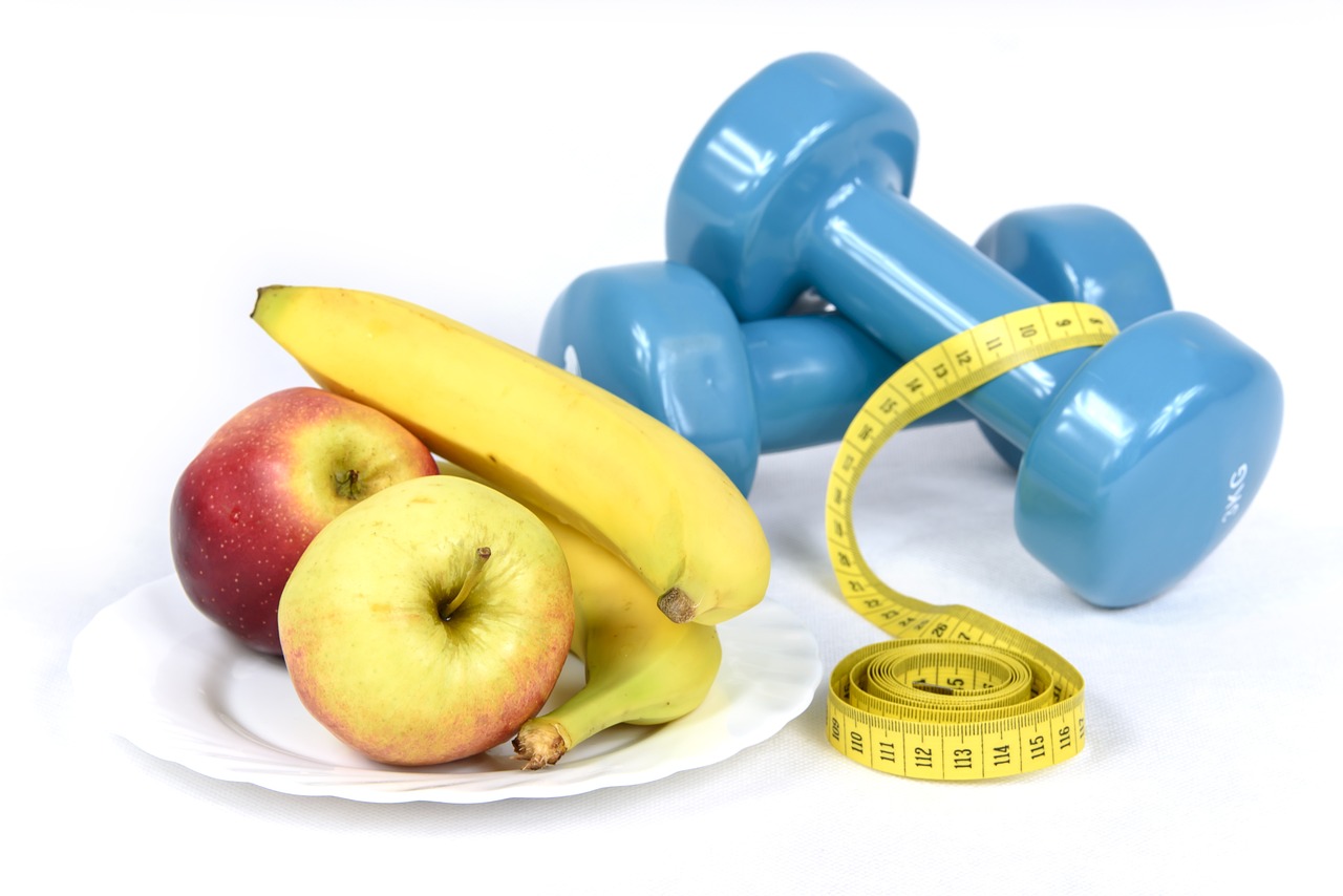 Dumbbells, apples, bananas, and a measuring tape on a white surface.